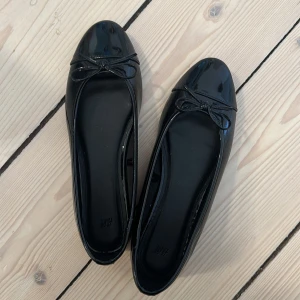 Svarta ballerinaskor från H&M - Glansiga svarta ballerinaskor från H&M med rund tå och söt rosettdetalj framtill. Använda endast en gång så är i väldigt fint skick! 