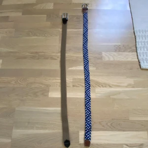 Två flätade bälten i brunt och blått - Säljer två snygga flätade bälten. Ett är helbrunt med svarta skinn-detaljer och silverspänne, det andra är blått med vitt och svart mönster samt bruna skinn-detaljer och silverspänne. Perfekta för att ge outfiten lite extra edge. De blå är 100cm och de bruna är 90cm.