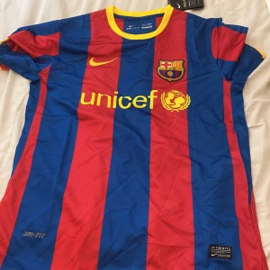FC Barcelona Nike fotbollströja - Klassisk FC Barcelona matchtröja från Nike med breda blå och röda ränder, gul Nike-logga och Unicef-tryck på bröstet. Tröjan har klubbmärket på vänster sida och är tillverkad i lätt Dri-FIT-material. Rund gul krage och korta ärmar.