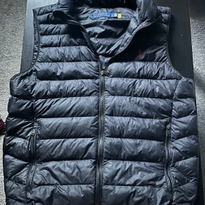 Svart dunväst från Polo Ralph Lauren - Snygg svart dunväst från Polo Ralph Lauren med horisontella quiltade paneler och dragkedja framtill. Västen har två praktiska fickor med dragkedja och en hög krage. Perfekt för lager-på-lager och stilren look.