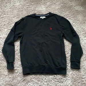 Svart sweatshirt från U.S. Polo Assn. - Svart sweatshirt från U.S. Polo Assn. med rund halsringning och röd broderad logga på bröstet. Tröjan har långa ärmar, ribbade muddar och är gjord i mjuk bomull. Enkel och stilren design som passar till det mesta. Pris går att diskutera 