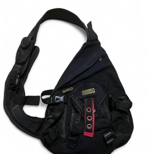 Svart axelväska från Diesel - Cool svart axelväska från Diesel med bred rem, flera fickor och detaljer i rött och silver. Väskan har spännen, dragkedja och en unik design med metallplatta och öljett-detaljer. Perfekt för dig som vill ha en streetstyle-vibe.