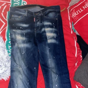 Dsquared2 blå jeans med färgstänk - Säljer ett par blå jeans från Dsquared2 med coola slitningar och vita färgstänk för en edgy vibe. Jeansen har klassisk femficksmodell, normal passform och tydliga kontrastsömmar. Perfekta för dig som vill sticka ut med din stil.