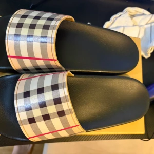 Burberry sandaler med klassiskt rutmönster - Snygga sandaler från Burberry med det ikoniska rutmönstret i beige, svart och rött på remmen. Svart platt sula i syntetmaterial, perfekt för sommaren. Stilsäkra slides som ger en lyxig touch till din outfit.