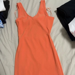 Orange ribbad kortklänning med knyt - Superfin orange kortklänning i ribbat material med v-ringning och ärmlös design. Baksidan har en snygg knytdetalj och öppen rygg. Perfekt för varma dagar när du vill sticka ut lite extra. I storlek S. Klänningen slutar vid knäna