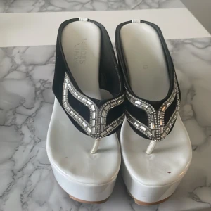 Svarta och vita platåsandaler med strass - Unika sandaler med hög vit platåsula och svarta remmar dekorerade med glittrande strass. Tåremmen är vit och sandalerna har öppen tå. Perfekta för dig som vill sticka ut med en glamorös och trendig look.