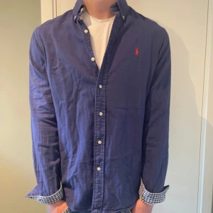 Ralph Lauren Skjorta - Snygg blå skjorta från Polo Ralph Lauren med klassisk krage och knappar hela vägen. Custom fit-modell med rutigt tyg på insidan av manschetterna och röd broderad logga på bröstet. Perfekt för dig som gillar stilrena plagg med en twist.
