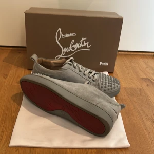 Grå Louboutins i mocka med nitar - Säljer ett par grå sneakers i mocka med coola nitar framtill och snörning. Skorna har en rund tå och platt sula, samt en röd undersida som ger en snygg kontrast. Perfekta för dig som vill sticka ut med din stil. Pris kan diskuteras.