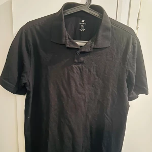 Svart pikétröja från H&M - Klassisk svart pikétröja från H&M i regular fit. Tröjan har korta ärmar, mjuk krage och knäppning framtill. Tillverkad i bomull för en skön känsla och enkel stil. Perfekt till jeans eller shorts för en clean look.