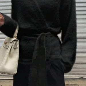 Svart fluffig omlottkofta med bälte - Svart, supermjuk och fluffig omlottkofta med bälte i midjan. Koftan har lång ärm, v-ringning och en loose passform som ger en chill och trendig vibe. Perfekt för lager-på-lager och att styla med smycken eller väska.