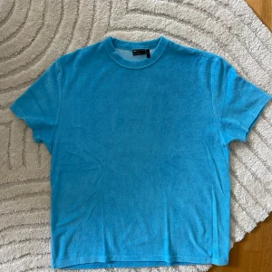 Blå frotté t-shirt från ASOS DESIGN - Snygg blå t-shirt från ASOS DESIGN i frottéliknande material. T-shirten har rund hals, korta ärmar och en relaxed fit. Perfekt för dig som vill ha en chill och unik vibe i garderoben. Helt ny och oanvänd!