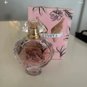 Olympea Blossom parfym Paco Rabanne - Olympea Blossom från Paco Rabanne är en lyxig parfym i en rund, genomskinlig glasflaska med guldiga detaljer och blommigt mönster. Doften kommer i en rosa ask med guldvingar och svarta blommor. Parfymen har en feminin och fräsch känsla med blommiga inslag. Har endast testat doften , 