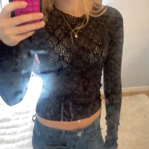 Svart spetsig långärmad topp - Säljer en svart, tight långärmad topp i spets med transparenta partier och rund hals. Sitter snyggt croppad och har ett mönster i spetsen som ger en cool och trendig vibe. Perfekt att styla med jeans eller kjol för en edgy look.