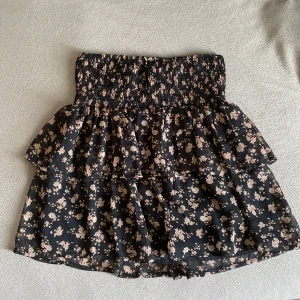 Blommig volangkjol från Vero Moda - Supersöt kort kjol från Vero Moda i svart med små beige blommor. Kjolen har smock i midjan och dubbla volanger som ger en flowy känsla. Perfekt för dig som gillar romantisk och trendig stil. Lätt och luftig, passar till sneakers eller boots.💕💕 andvänd bara en gång