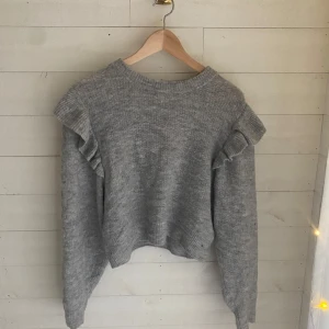 Grå stickad tröja med volangdetalj - Supermjuk grå stickad tröja med volangdetaljer på axlarna. Tröjan har en något croppad passform. 