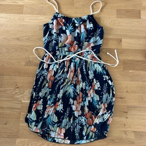 Blommig sommarklänning Holly Bracken - Supersöt kortklänning från Holly Bracken med marinblå botten och färgglatt blommönster. Klänningen har smala axelband, knyt i midjan och är tillverkad i ett lätt, luftigt material. Perfekt för varma dagar när du vill känna dig både chill och snygg.