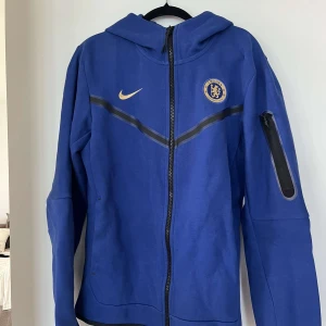 Chelsea Nike blå hoodiejacka S - Blå hoodiejacka från Nike med Chelsea FC-logga på bröstet och guldfärgad Nike-swoosh. Jackan har hel dragkedja, huva och en praktisk ficka med dragkedja på ärmen. Perfekt för dig som älskar fotboll och vill representera Chelsea.