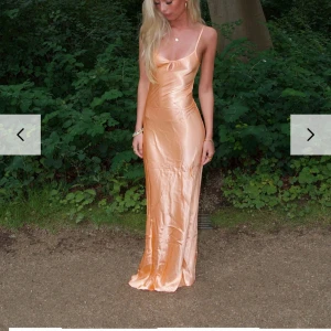 Champagnefärgad långklänning i satin - Säljer en champagnefärgad långklänning i glansigt satinmaterial. Klänningen har smala axelband, djup urringning och öppen rygg med knyt. Sitter figurnära och har en elegant, slinky siluett. Perfekt för dig som vill sticka ut med en lyxig vibe. XS/S