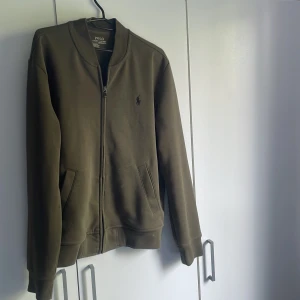 Olivgrön zip från Polo Ralph Lauren - Snygg olivgrön zip från Polo Ralph Lauren med dragkedja framtill och två snedställda fickor. Jackan har klassisk ribbad krage och muddar, samt diskret broderad logga på bröstet. Perfekt för dig som gillar stilren och sportig look.