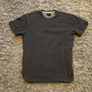 Mörkgrå t-shirt från DC, storlek M - En stilren mörkgrå t-shirt från DC i storlek M. T-shirten har klassisk rund halsringning, korta ärmar och en liten DC-logga nertill. Tillverkad i mjuk bomull för skön känsla och enkel stil.