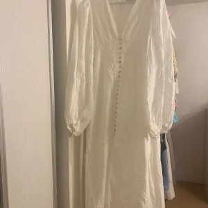 Adoore positano 34 - Superfin vit långklänning från Adoore med v-ringning, långa puffärmar och dekorativa knappar framtill. Klänningen har broderade detaljer i tyget och dragkedja bak. Perfekt för sommarens speciella tillfällen.
