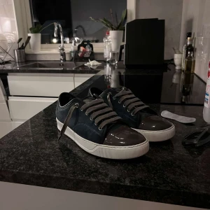 Lanvin sneakers i blå mocka och lack - Snygga sneakers från Lanvin i mörkblå mocka med grå lackad tå och grå snörning. Skorna har vit platt sula och rund tå, vilket ger en clean och modern look. Perfekta för dig som gillar exklusiva detaljer och stilren design. Med frågor kan ni Bara skriva til mig 