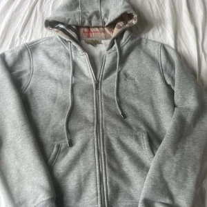 Grå hoodie från Burberry med dragkedja - Snygg ljusgrå hoodie från Burberry med klassiskt rutigt foder i huvan och broderad logga på bröstet. Tröjan har dragkedja, snörning i huvan och två fickor framtill. Perfekt för dig som gillar stilrena och exklusiva plagg.