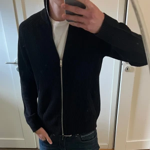 Svart ribbad kofta från Jack & Jones - Säljer en svart ribbstickad kofta från Jack & Jones med ful zip. Den är använd en gång och har legat i en låda sedan dess. Säljer på grund av att en polare har en likadan så vill inte ha den. 