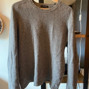 Grå stickad tröja från Paul & Friends - En klassisk grå stickad tröja från Paul & Friends i storlek L. Tröjan har rund halsringning, ribbade muddar och är långärmad. Perfekt för lager-på-lager och funkar till både jeans och chinos. Enkel och stilren design som passar till det mesta.