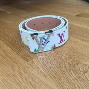 Vit Louis Vuitton bälte med färgglada loggor - Snyggt vitt bälte från Louis Vuitton med ikoniska LV-loggor i flera färger och silverfärgat LV-spänne. Bältet är gjort i skinn och har en clean look med färgglada detaljer som sticker ut. Perfekt accessoar för att lyfta din outfit. Har kvitto på den och kort.