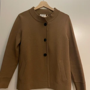 Beige cardigan jacka från Isay - Stilren beige cardigan jacka från Isay med svarta knappar framtill och två fickor. Jackan har en rak passform, lång ärm och en snygg detalj i ryggen. Perfekt för lager-på-lager och enkel att matcha med olika outfits.