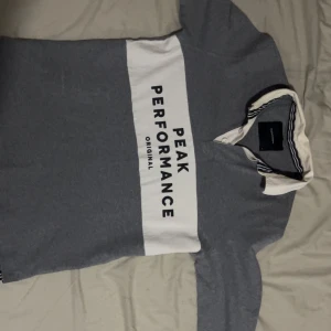 Grå pikétröja Peak Performance M - Snygg grå långärmad pikétröja från Peak Performance med vit krage och bred vit rand över bröstet där loggan står tryckt. Tröjan har knappar vid halsen och coola svarta ränder på insidan av kragen. Perfekt för en sportig och stilren look.