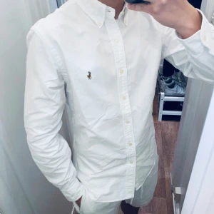 Vit skjorta från Polo Ralph Lauren - Klassisk vit skjorta från Polo Ralph Lauren i slim fit-modell. Skjortan har långa ärmar, knappar framtill och den ikoniska broderade loggan på bröstet. Perfekt för en clean och stilren look. (Original pris: 2095kr)