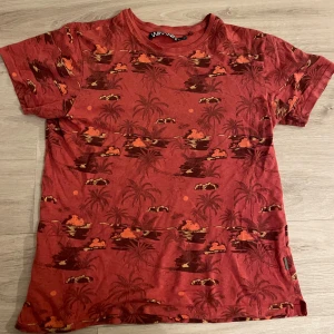 Röd t-shirt med tropiskt mönster Win Win - Snygg röd t-shirt från Win Win med tropiskt print av palmer, grottor och solnedgångar i mörkare toner. T-shirten har rund hals, korta ärmar och är gjord i mjukt material som känns skönt mot huden. Perfekt för dig som gillar färg och unika prints.