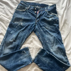Dsquared2 blå jeans med slitningar - Snygga blå jeans från Dsquared2 med coola slitningar och färgstänk för en edgy look. Klassisk femficksmodell med raka ben och knappgylf. Tydliga Dsquared2-detaljer och röd logotagg vid gylfen. Perfekta för dig som gillar streetstyle. 100% äkta från NK 