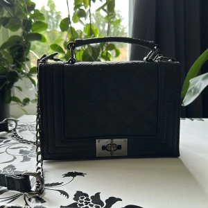 Svart axelväska med kedja och ormmönster - Stilren svart axelväska med ormmönstrad struktur och silverfärgad kedjerem. Väskan har ett topphandtag, rektangulär form och snyggt vridlås framtill. Perfekt storlek för det viktigaste och ger en edgy vibe till din outfit.