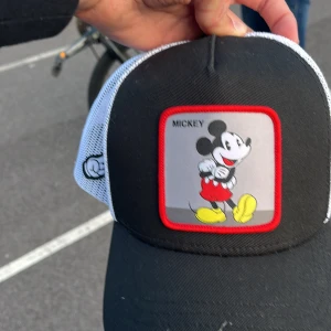 Capslab keps med Mickey Mouse - Snygg svart och vit keps från Capslab med mesh på sidorna och bak, justerbar snapback. Framtill finns en stor patch med Mickey Mouse i rött, gult och grått, inramad med röd kant. Cool detalj med Mickeys hand broderad på sidan.