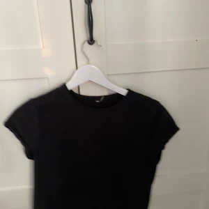 Svart croppad t-shirt - En enkel svart croppad t-shirt med rund hals och korta ärmar. Perfekt att matcha med jeans eller kjol för en clean look. Mjuk och skön bomullskänsla.