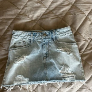 Ljusblå jeanskjol med slitningar - Snygg ljusblå jeanskjol med slitna detaljer och fransig nederkant. Klassisk femficksmodell med bälteshällor och dragkedja fram. Perfekt för dig som gillar en avslappnad och trendig stil.