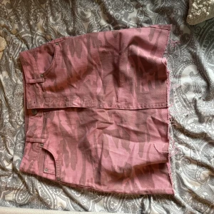 Rosa camo jeanskjol med rå kant - Säljer en rosa kort jeanskjol med camouflagemönster och rå, fransig kant nertill. Kjolen har klassiska jeansdetaljer som fickor och knappstängning framtill. Storlek 36 men skulle säga 34