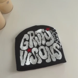 Svart mössa med tryck  - Cool svart mössa med vit grafisk text, röda stjärnor och ett snyggt spindelnätsmönster på sidan. Perfekt accessoar för dig som gillar streetstyle och vill sticka ut. Mjuk och stretchig, passar de flesta huvuden.