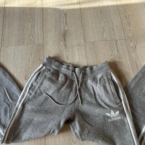 Grå mjukisbyxor från Adidas - Säljer ett par grå mjukisbyxor från Adidas med klassiska vita ränder längs sidorna. Byxorna har en bekväm resårmidja med snörning och sidofickor. Perfekta för en avslappnad stil.