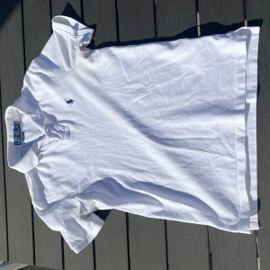 Vit pikétröja från Ralph Lauren - Säljer en klassisk vit pikétröja från Ralph Lauren med broderad logga i blått på bröstet. Tröjan har korta ärmar och en stilren krage med knappar. Perfekt för en avslappnad och stilren look.