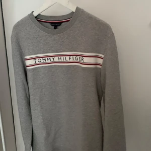 Tommy Hilfiger grå sweatshirt, storlek M - Grå sweatshirt från Tommy Hilfiger med märkets namn tryckt över bröstet. En klassisk och bekväm tröja som passar perfekt till vardags.