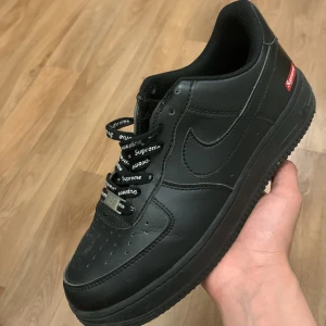 Svarta Nike Air Force x Supreme - Säljer ett par svarta Nike Air Force i samarbete med Supreme. Skorna har en klassisk design med Supreme-logga på sidan och snören med Supreme-text. Perfekta för streetwear-entusiaster!
