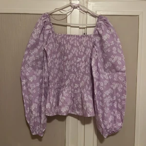 Lila blommig topp från Pernilla Wahlgren - Säljer en söt lila topp med vita blommor från Pernilla Wahlgrens kollektion. Toppen har långa puffärmar och sitter fint på, men inte min stil och aldrig använd! Nypris: ≈299kr