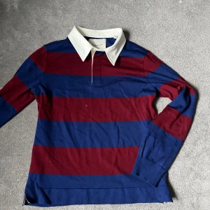 Randig pikétröja från GANT Merinoull  - Nypris 1500kr. Säljer en snygg randig pikétröja från GANT i marinblått och vinrött bara för 500kr. Kan gå ner i pris. Tröjan är tillverkad i extra fin merinoull och är riktigt skön. Knappt använd. Köpt på NK. Perfekt för en unik och laidback look. Storlek M.  