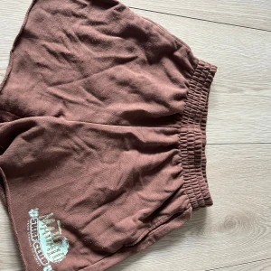 Bruna shorts från Divided - Säljer ett par bruna shorts från Divided med elastisk midja och tryck av 'Maui Surf Club' i vitt och grönt. Perfekta för en avslappnad dag på stranden eller hemma. 🏄‍♀️🩷