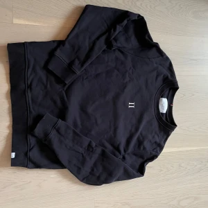 Svart sweatshirt med logotyp - Säljer en stilren svart sweatshirt med en liten logotyp på bröstet. Tröjan har långa ärmar och ribbade muddar vid ärmslut och nederkant. Perfekt för en avslappnad look.