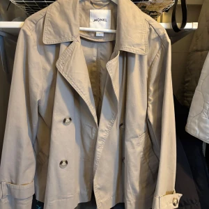 Beige trenchcoat från Monki - Säljer en stilren beige trenchcoat från Monki. Jackan har en klassisk krage och knappar framtill. Den är långärmad med justerbara ärmslut och har en rak passform. Perfekt för vår och höst!